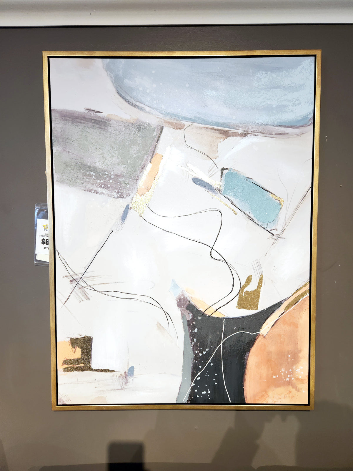 The Laurent Framed Canvas by Crestview Collection showcases an abstract design with vibrant mustard and blue hues, complemented by striking lines and shapes. It features soft colors such as white, gray, and accents of gold, all elegantly framed in a gold frame on the wall.