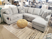 The Decor-rest 2541 Sectional, featuring Canadian craftsmanship, steals the spotlight with its gray upholstery. It's paired with patterned pillows and accompanied by a round wicker ottoman on a beige rug, all highlighting minimalist styling in a chic furniture store setting.