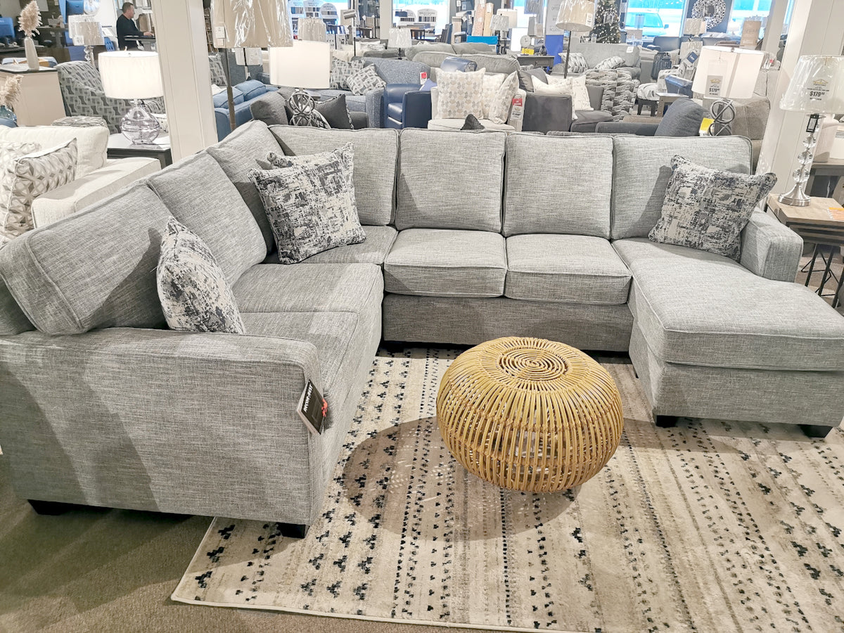 The 2541 Sectional by Decor-rest, upholstered with luxurious Canadian fabric, includes patterned throw pillows and a round wicker ottoman on a patterned rug, all showcasing minimalist styling in the furniture store.