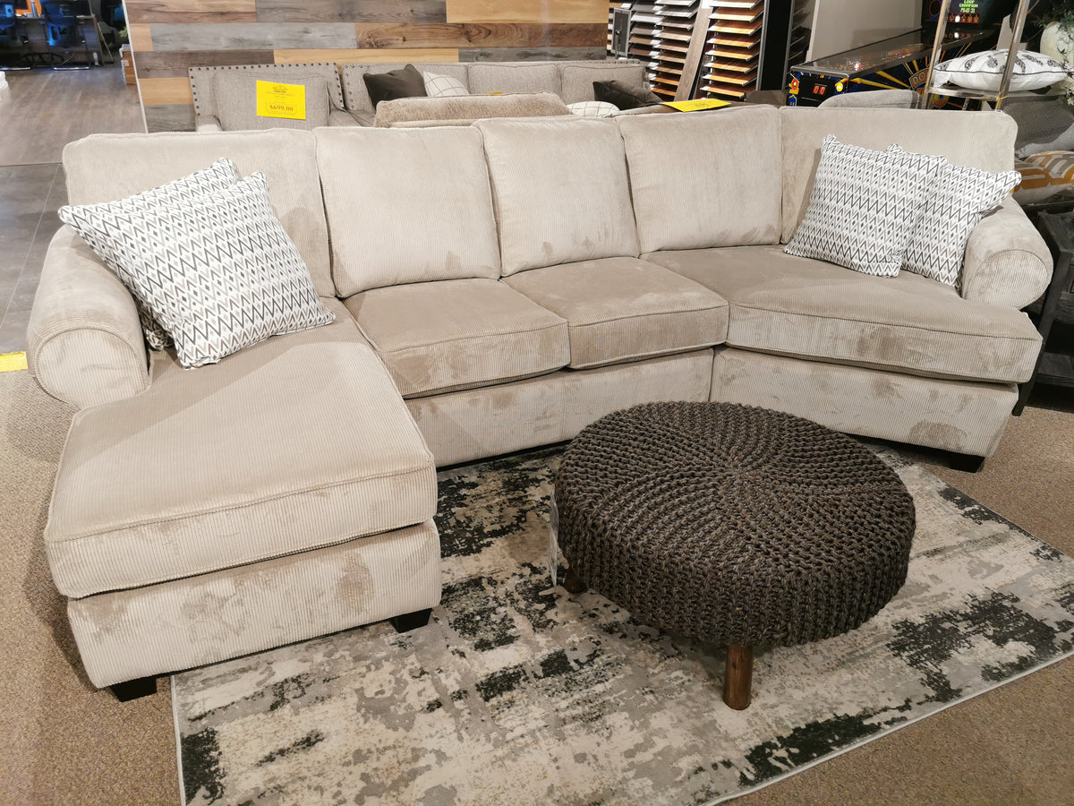 The Decor-rest 2566/83 Sectional, featuring rolled arms and patterned pillows, is displayed alongside a round, dark knitted ottoman on a patterned rug in the showroom, highlighting the exceptional craftsmanship of hand-built upholstery artisans.