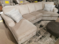 The 2566/83 Sectional by Decor-rest, a beige traditional piece with rolled arms, is accented with two patterned cushions atop a rug. A round, dark ottoman adds an elegant touch to the setup.