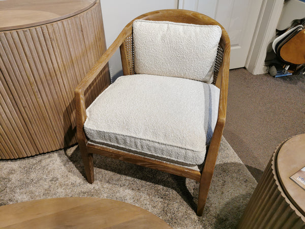 The Landon Accent Chair by Mercana, featuring white cushions, sits stylishly on a carpeted floor near round wooden furniture and a guitar case. It's an elegant touch that any interior expert would appreciate.