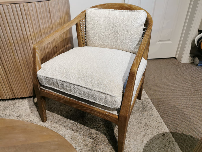 A stylish and elegant interior showcases the Mercana Landon Accent Chair, featuring a wooden frame with a cushioned seat and backrest, gracefully positioned on a carpeted floor beside a round table.