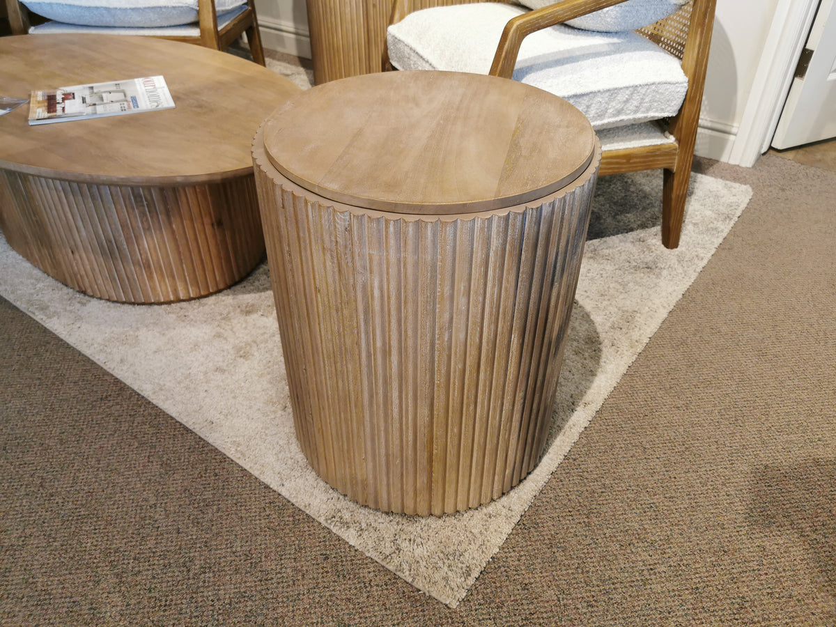 The 69882 Terra Round Side Table by Mercana, crafted from solid mango wood, stands elegantly on a carpet beside chairs and a coffee table in the living room, showcasing its ribbed cylindrical design.