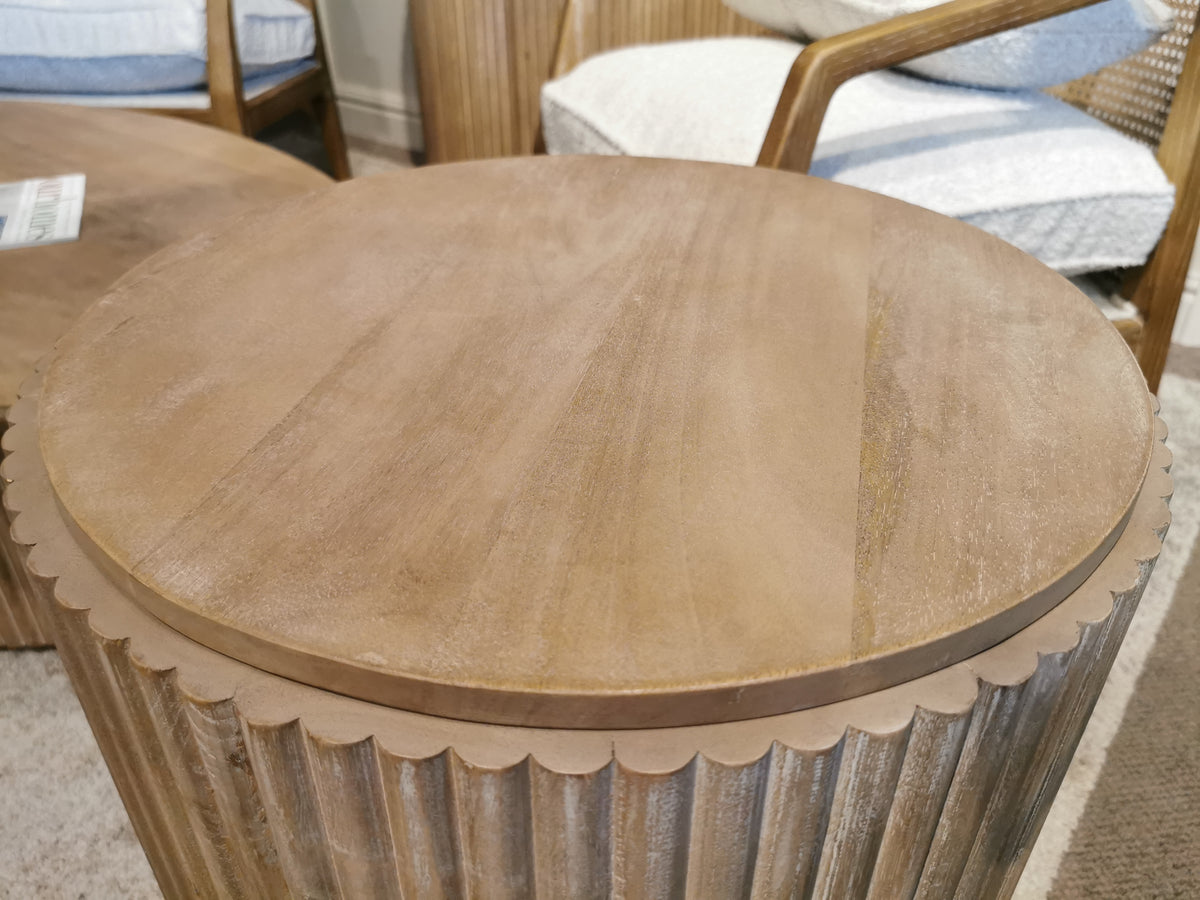 A round Terra Side Table from Mercana, identified as product 69882 and crafted from solid mango wood with a textured edge, is placed next to a cushioned wooden chair on a carpeted floor.