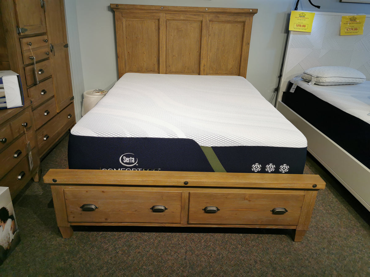 The B5487 Lynnfield Queen Bed by Magnussen offers rustic charm with its sturdy pine construction and two drawers in the footboard, perfectly paired with a Serta mattress. It is currently showcased in a furniture showroom.