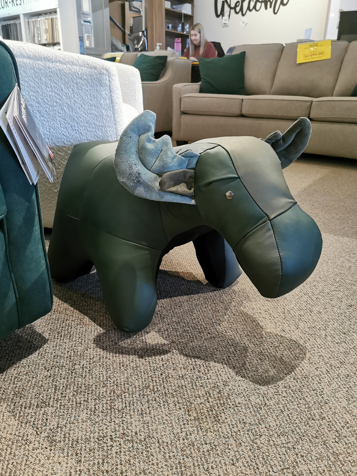 In a furniture showroom, Reiner's Originals' handcrafted green leather moose-shaped footrest from the Reiner's Leather Animal Ottomans collection sits charmingly on the carpeted floor—a unique piece in their animal ottomans range.