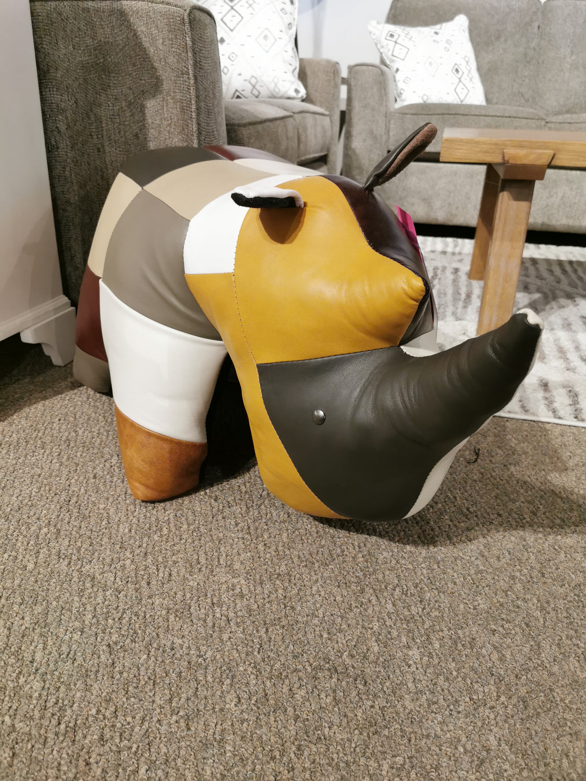 A multicolored, patchwork fabric rhino-shaped footrest from Reiner's Leather Animal Ottomans by Reiner's Originals graces the carpeted floor next to a wooden table, showcasing its handcrafted charm among other similar pieces.