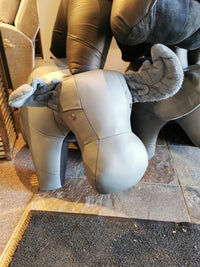 A gray, handcrafted moose-shaped footstool from Reiner's Originals, featuring fabric ears and tail and set on a stone floor in the store, embodies the charm of Reiner's Leather Animal Ottomans.