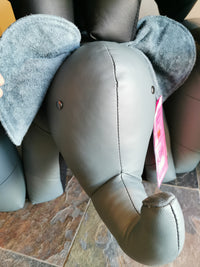 A close-up of a plush toy that resembles one of Reiner's Leather Animal Ottomans, featuring an elephant shape with large fabric ears and a tag on its trunk, is set on a tiled floor.