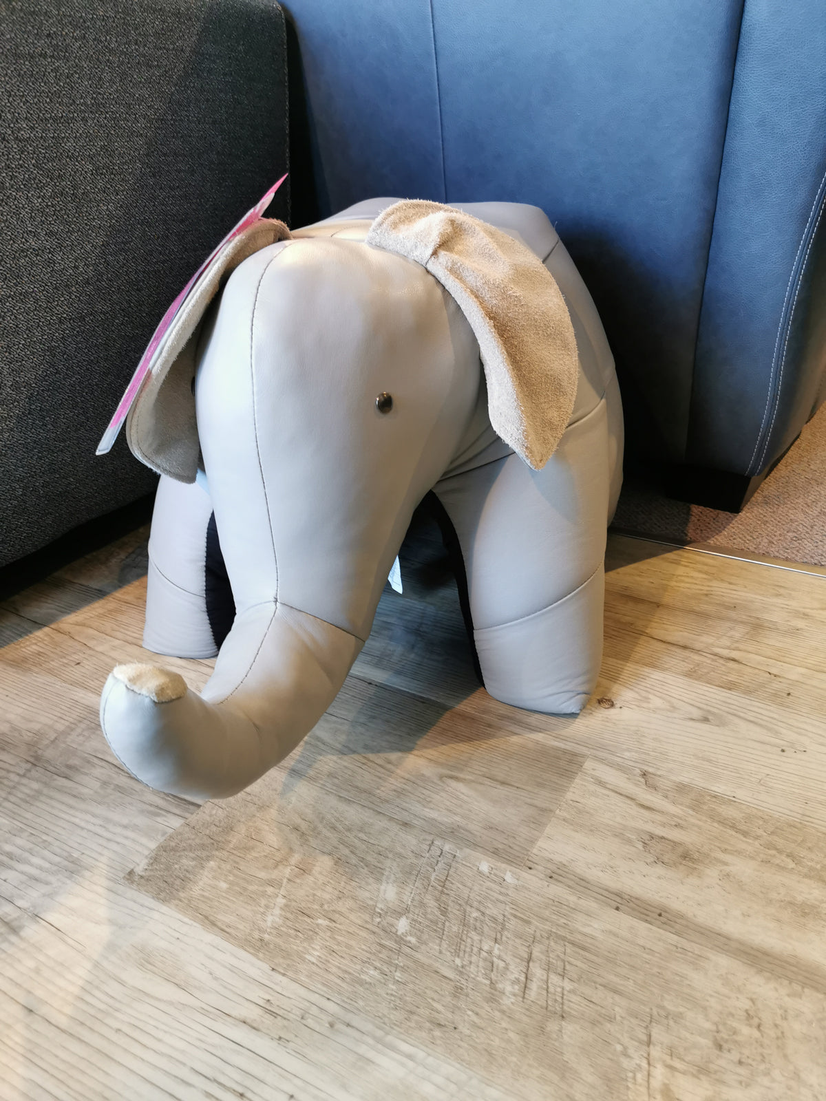 A beige stuffed elephant toy with floppy ears stands on a wooden floor, complementing the cozy charm of Reiner's Leather Animal Ottomans by Reiner's Originals near the sofa.