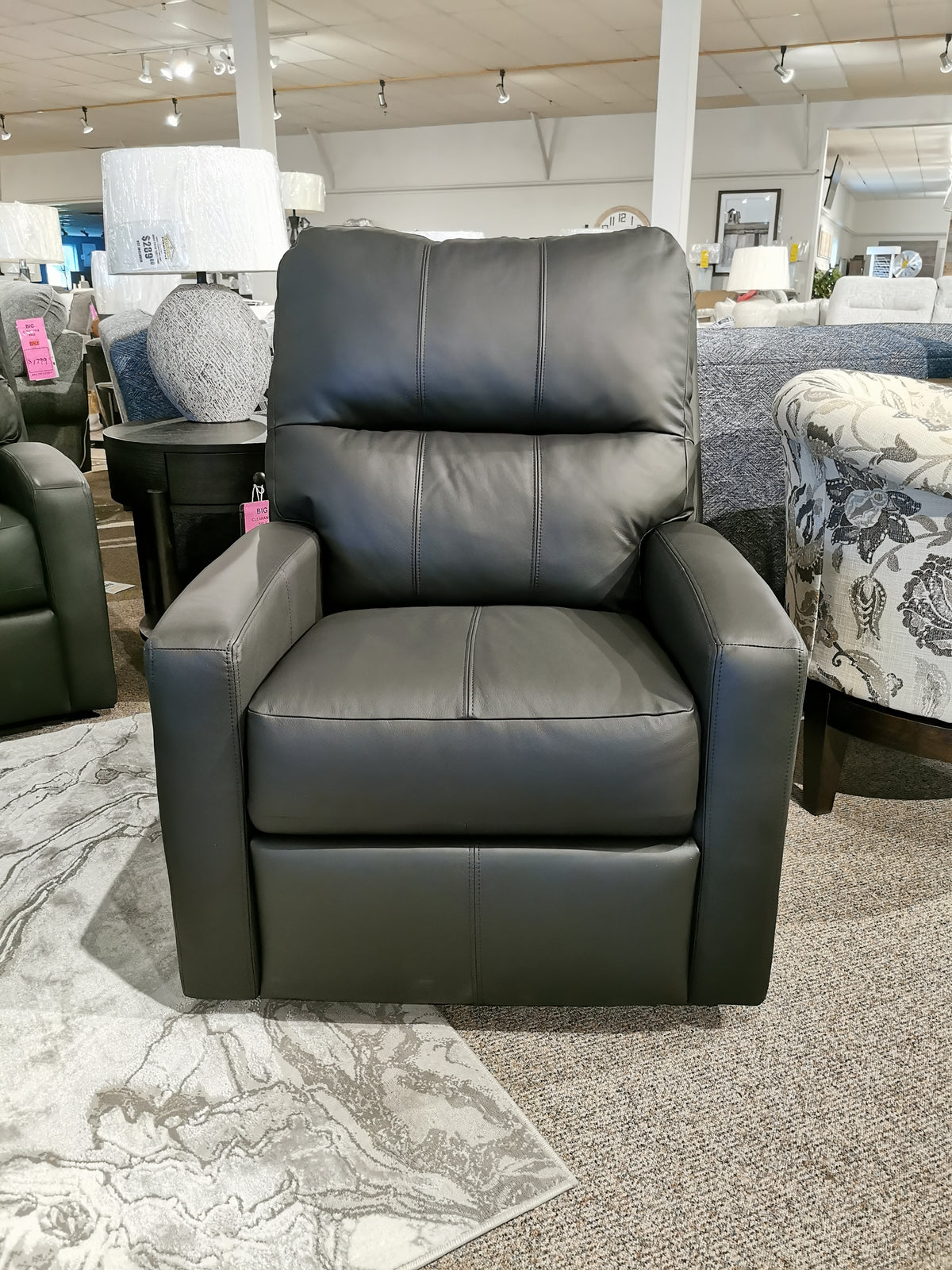 In the showroom, a black leather M38505 Power Reclining Chair by Decor-rest stands out, flaunting its sleek design alongside other furniture. This model offers a powered option with headrest and lumbar support, enhancing the appeal of our Power Reclining Loveseat collection.