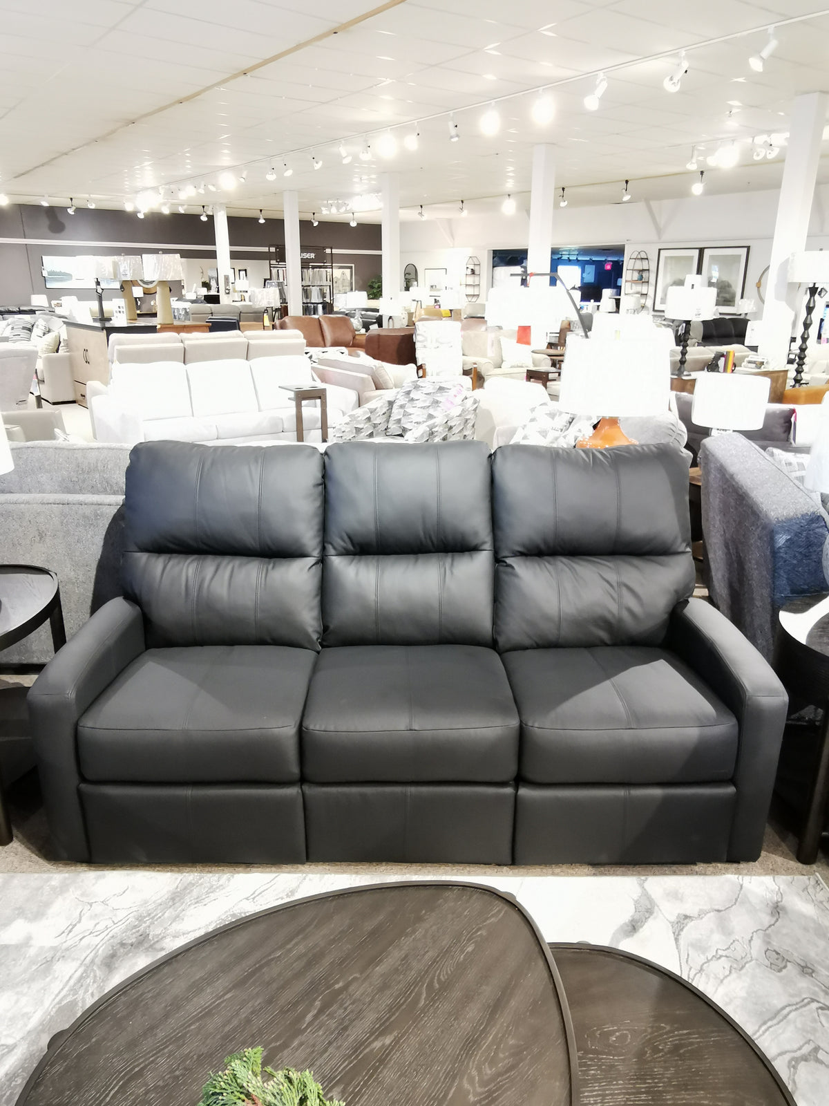 A Decor-rest M38505 power reclining sofa in black leather, featuring lumbar support, is showcased in a furniture showroom along with an assortment of other couches and tables.