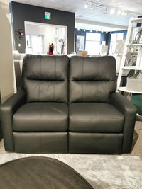 In the store showroom, the sophisticated Decor-rest M38505 Power Reclining Loveseat stands elegantly, framed by modern decor. This sleek black leather piece is enhanced with powered features, including an adjustable headrest and lumbar support, offering unmatched comfort and style.