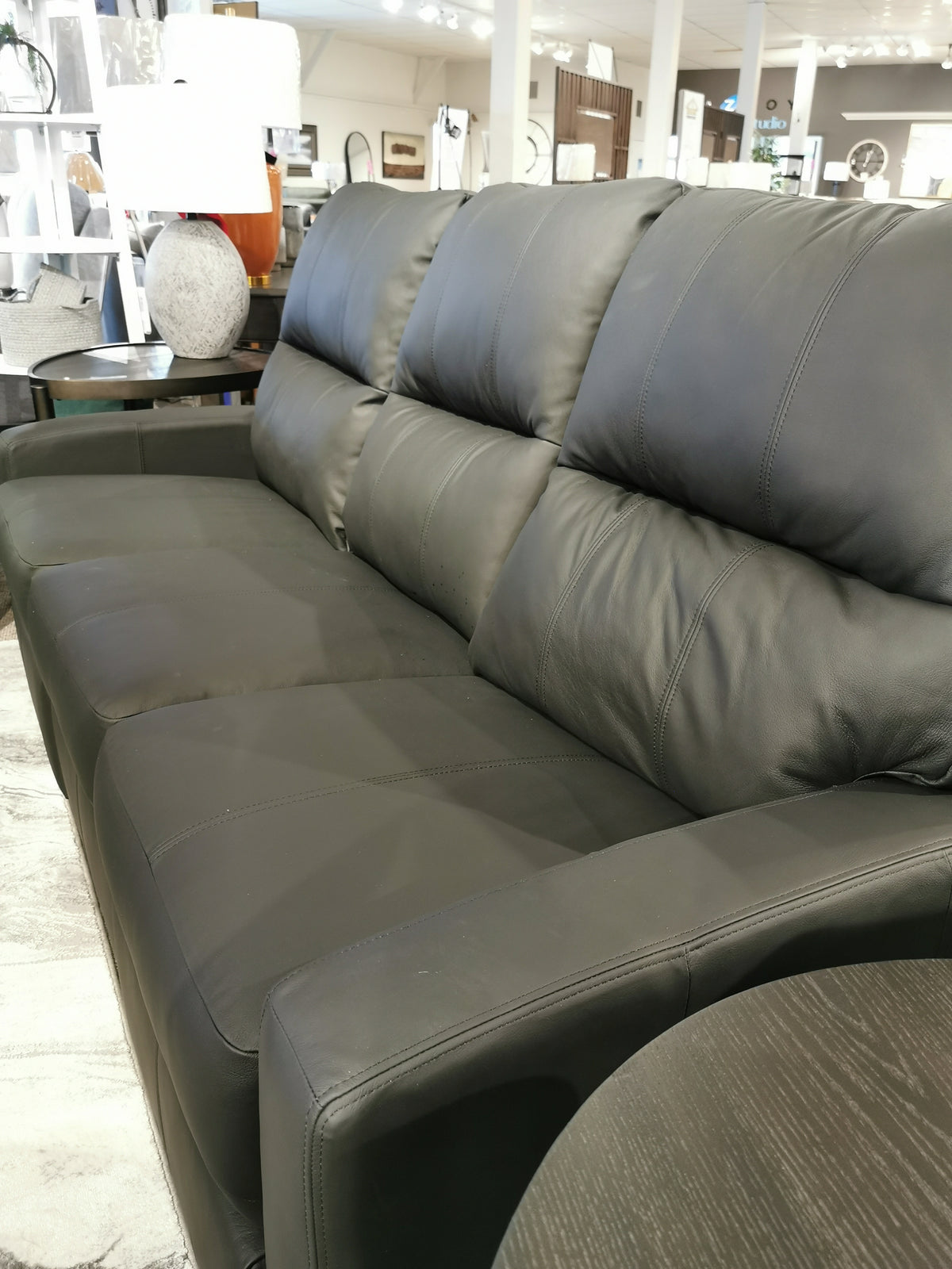 The Decor-rest M38505 Power Reclining Sofa in black leather, featuring padded cushions and lumbar support, is displayed in a well-lit showroom with chic side tables and elegant decor.