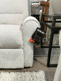 A light gray M8508 Power Reclining Chair by Decor-rest, featuring traditional arms and a tag attached to the armrest, sits elegantly next to a black metal side table on a carpeted floor.