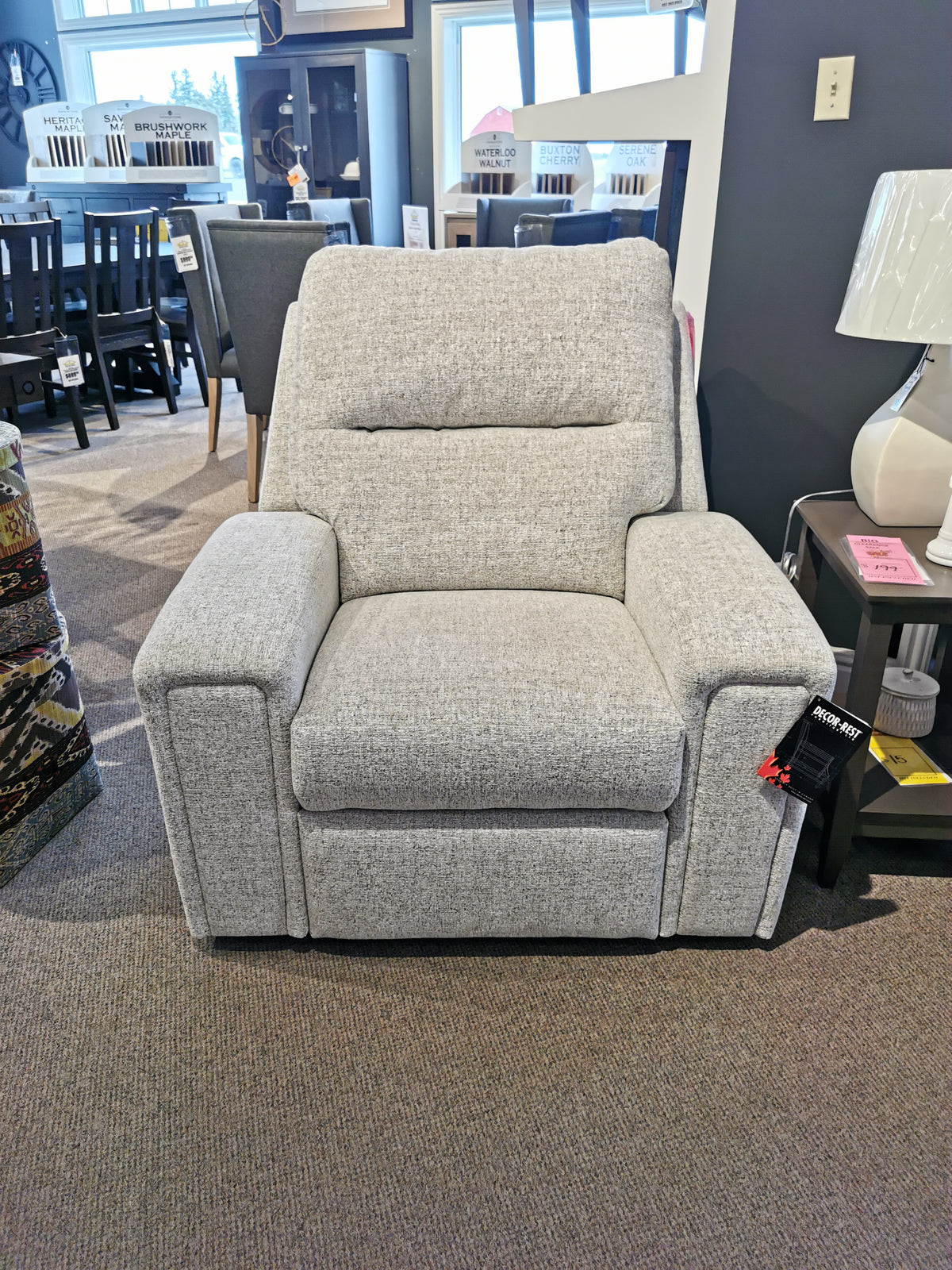 The M8500 Power Reclining Chair by Decor-rest, designed in Canada and featuring exquisite gray upholstery, is showcased in the furniture showroom with a lamp on a side table nearby.