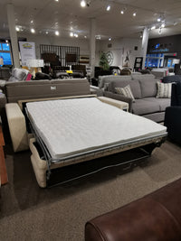 On display in the furniture store showroom is the Mathias Queen Sofa Bed by Donald Choi, featuring an extended queen foam mattress for demonstration. Amidst a selection of other sofas and seating options, this versatile piece offers high-density foam seats that guarantee both style and comfort.