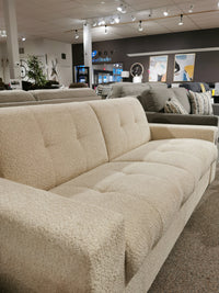 A beige couch with tufted cushions and high-density foam seats is displayed in a furniture showroom alongside other sofas, decorative items, and the versatile Mathias Queen Sofa Bed by Donald Choi.