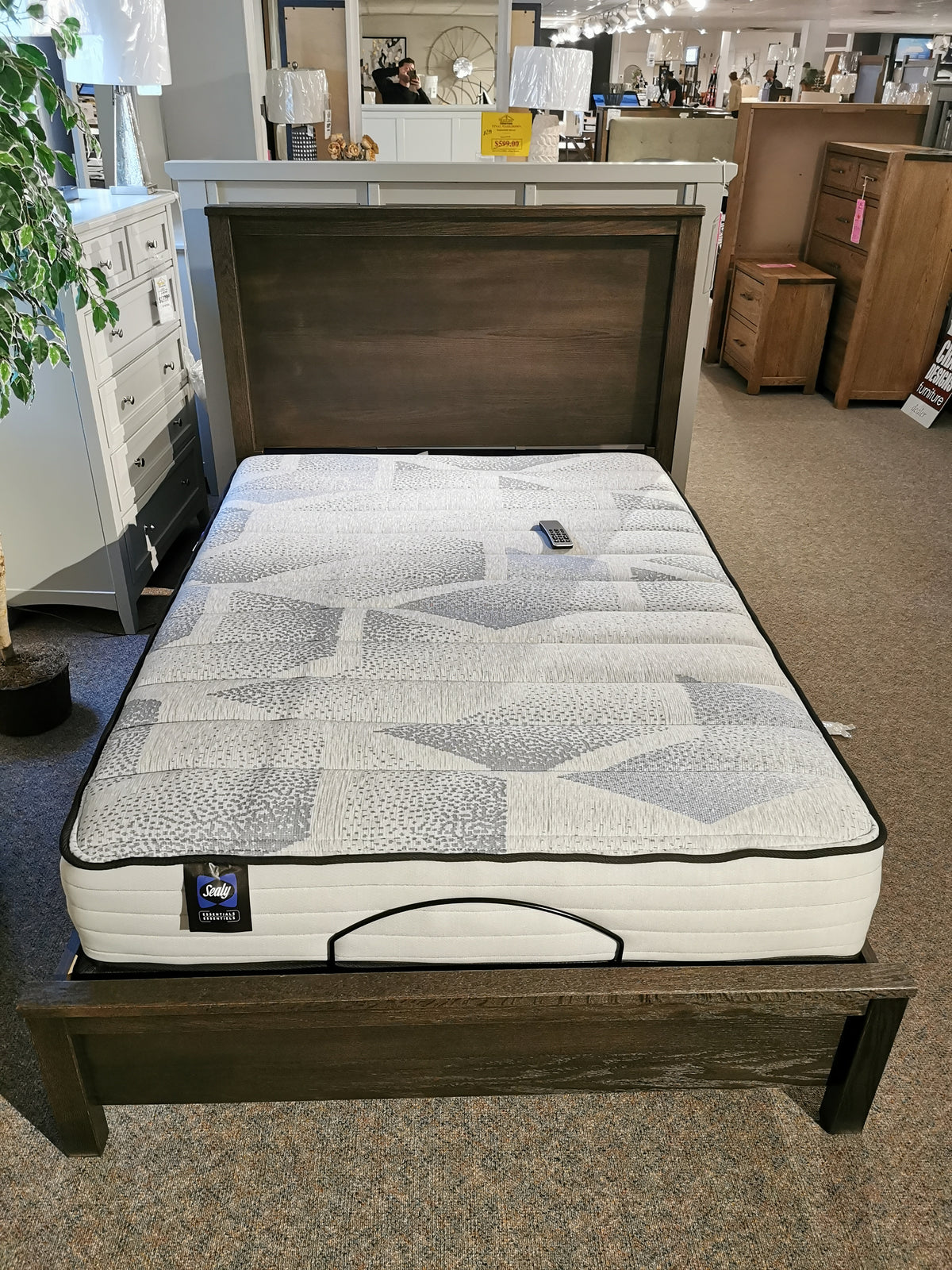 A display bed featuring a cushioned mattress sits in a furniture store setting, surrounded by an array of bedroom furniture pieces, including the Fundamental Panel Double Bed by Vaughan Bassett.