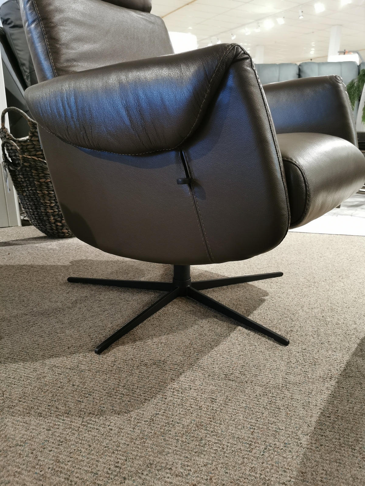 The Licia Recliner with Ottoman by Donald Choi, crafted from sleek black Italian leather and complemented by a metal base, elegantly rests on a beige carpet, offering a refined motion mechanism for ultimate comfort.