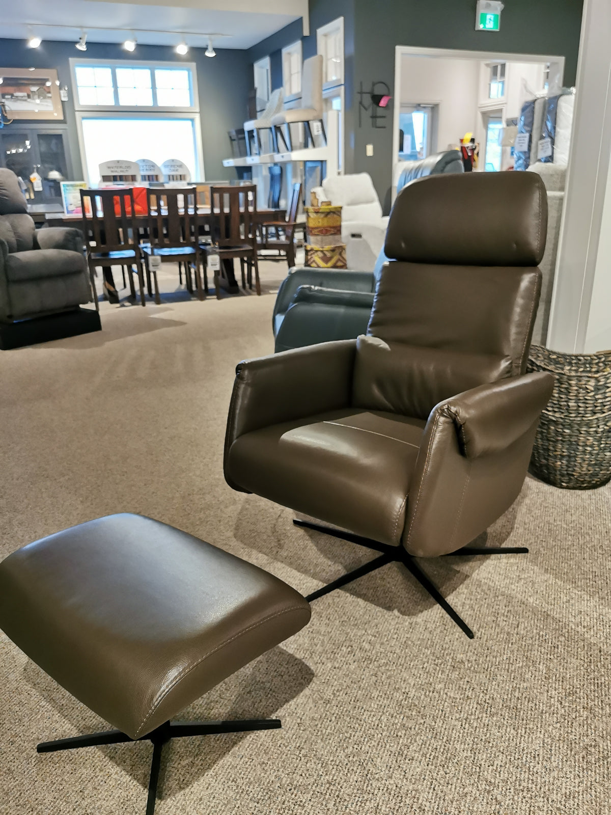 The Licia Recliner with Ottoman by Donald Choi, crafted from brown Italian leather and featuring a smooth motion mechanism for ultimate comfort, is on display in a furniture showroom. A stylish dining table set can be seen in the background.