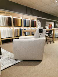 The showroom features fabric swatches on the wall and a luxurious 40111 Sanibel Power Wall Recliner by Palliser, equipped with heat and massage for an enhanced motion experience. A clock, tables, and chairs complete the inviting scene.