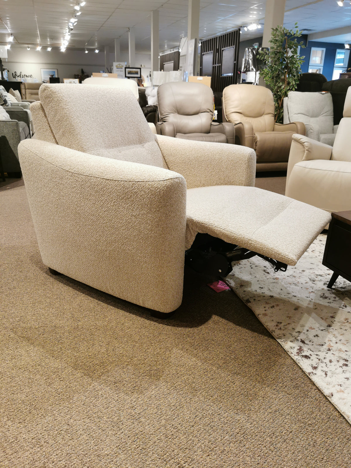 The 40111 Sanibel Power Wall Recliner by Palliser, featuring a beige upholstered design and an extended footrest, delivers a premium motion experience, complemented by the showroom's array of other chairs and a potted plant in the background.