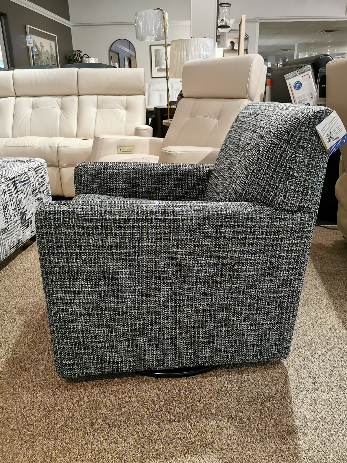 Side view of the Pia Swivel Chair in gray upholstery by Palliser, displayed in a furniture store on a brown carpet; its versatile design subtly complements the other furniture in the background.