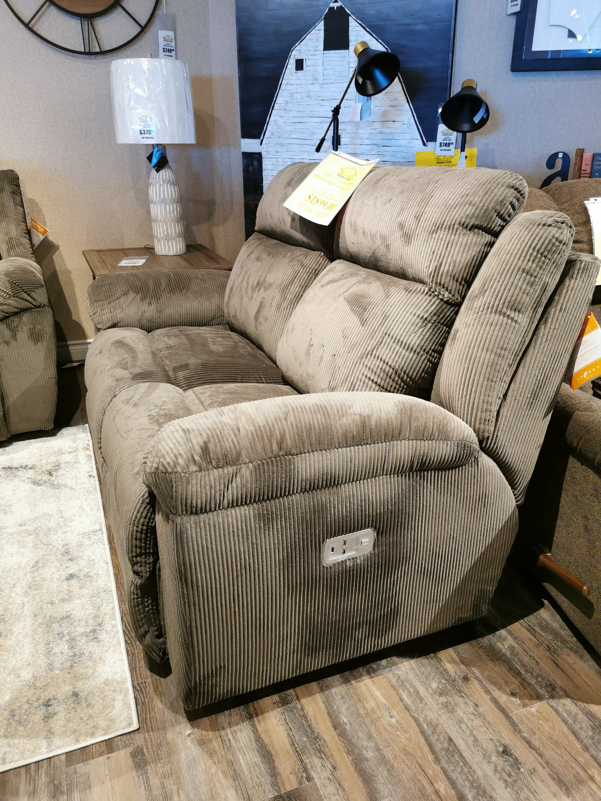 The 761 Joel Power Reclining Loveseat by La-Z-Boy, crafted in luxurious brown corduroy, is showcased in a showroom. It boasts electronic controls and cushioned arm pads. Complemented by a nearby lamp and striking wall art, it creates an inviting ambiance.