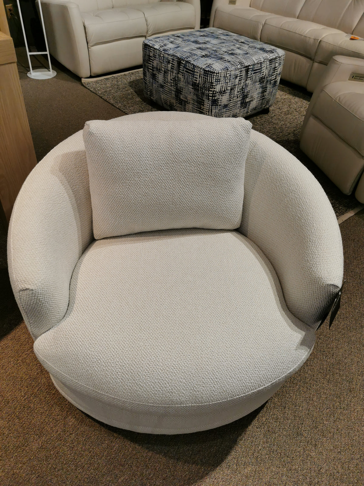 A fashionable accent piece, the Palliser Kinsley Swivel Chair showcases a circular, textured design with a complementary cushion. It is elegantly positioned on a carpeted floor beside a patterned ottoman and beige couches.