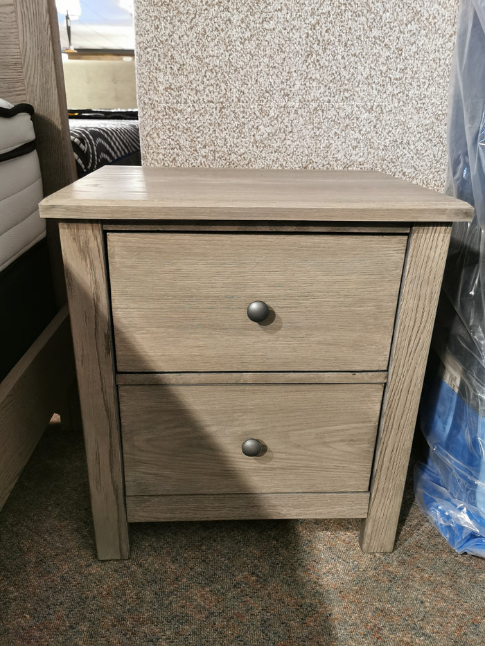 The Fundamentals 2-Drawer Nightstand by Vaughan Bassett, crafted from oak with a light wood finish, features two drawers accented with round metal knobs, and is set against a textured wall backdrop.