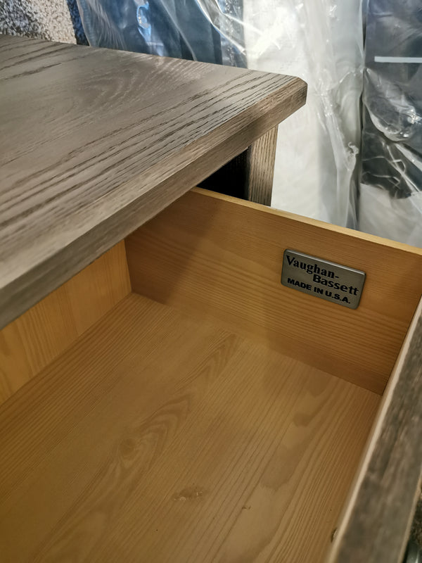 Open the drawer of a solid oak wooden cabinet to reveal a label inside that reads "Vaughan Bassett." In the background, a plastic-wrapped item suggests ample storage possibilities within this elegant Fundamentals 2-Drawer Nightstand.