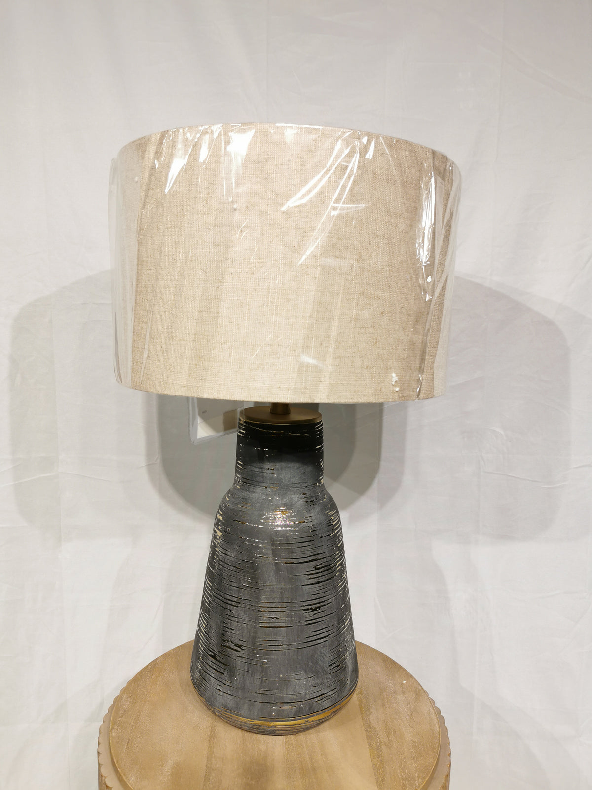 The Crestview Collection Trinity Table Lamp has a beige fabric shade, a textured charcoal ceramic base, and sits on wood.