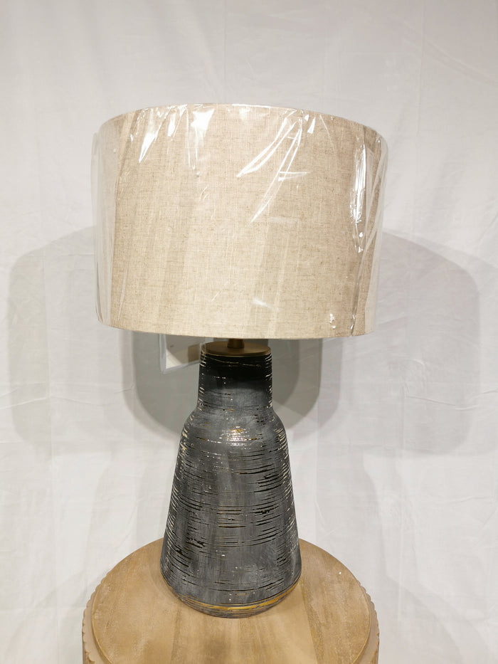 The Crestview Collection Trinity Table Lamp has a beige fabric shade, a textured charcoal ceramic base, and sits on wood.