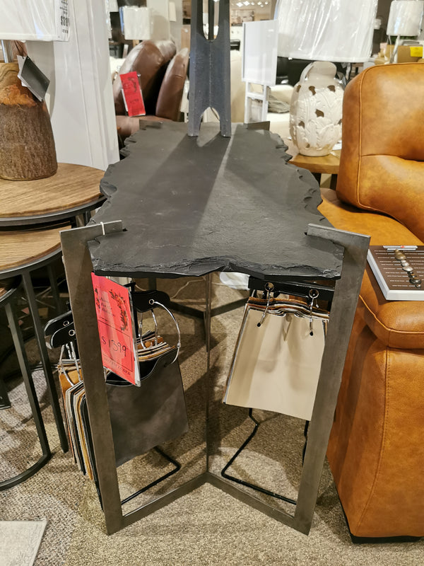 A Mercana 69050 Shale Sofa Table with a black live-edge slate top and double pedestal base in a furniture store.