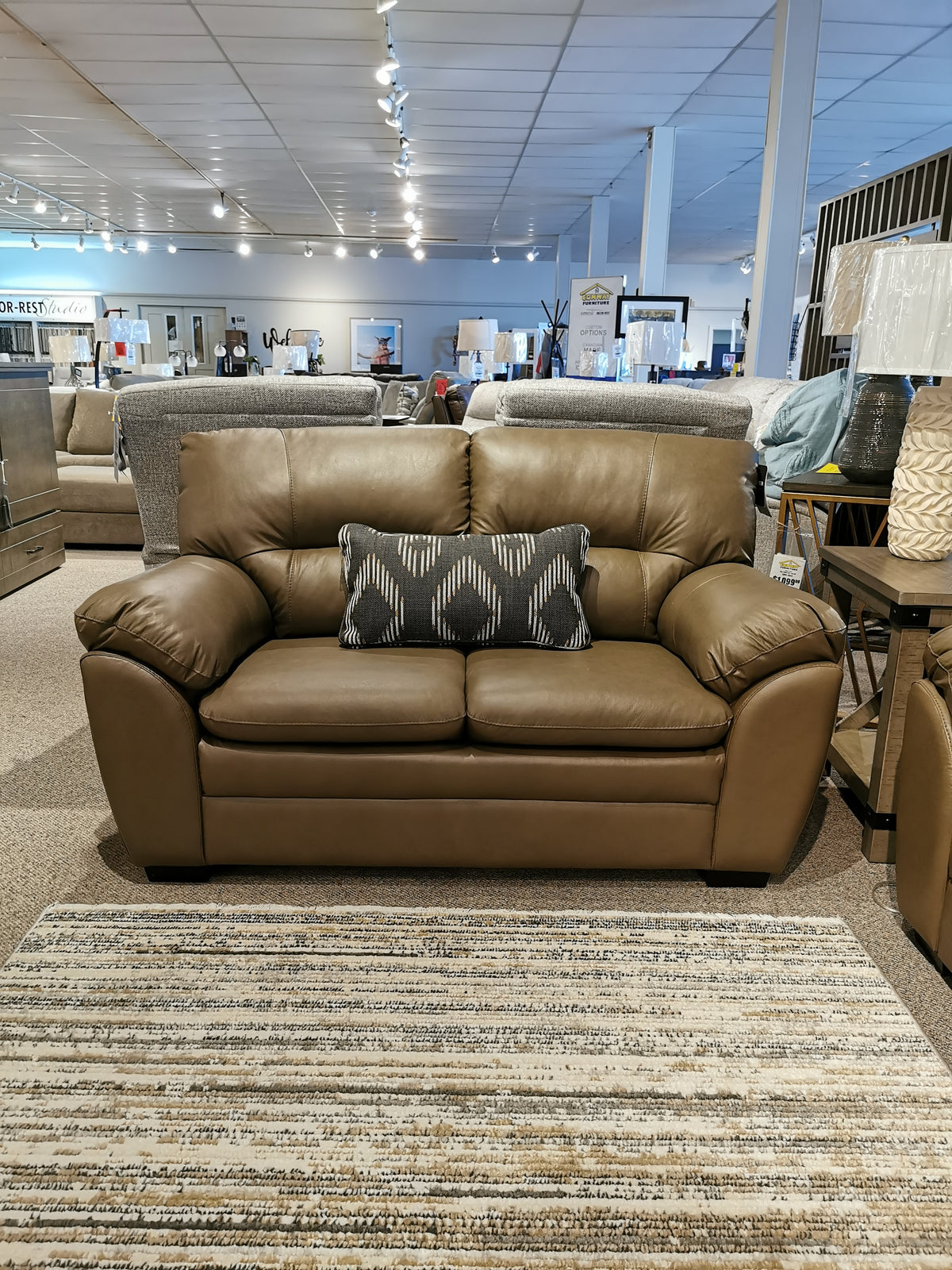 A Palliser 77343 Amisk leather loveseat with patterned cushion sits on a striped rug in the showroom.