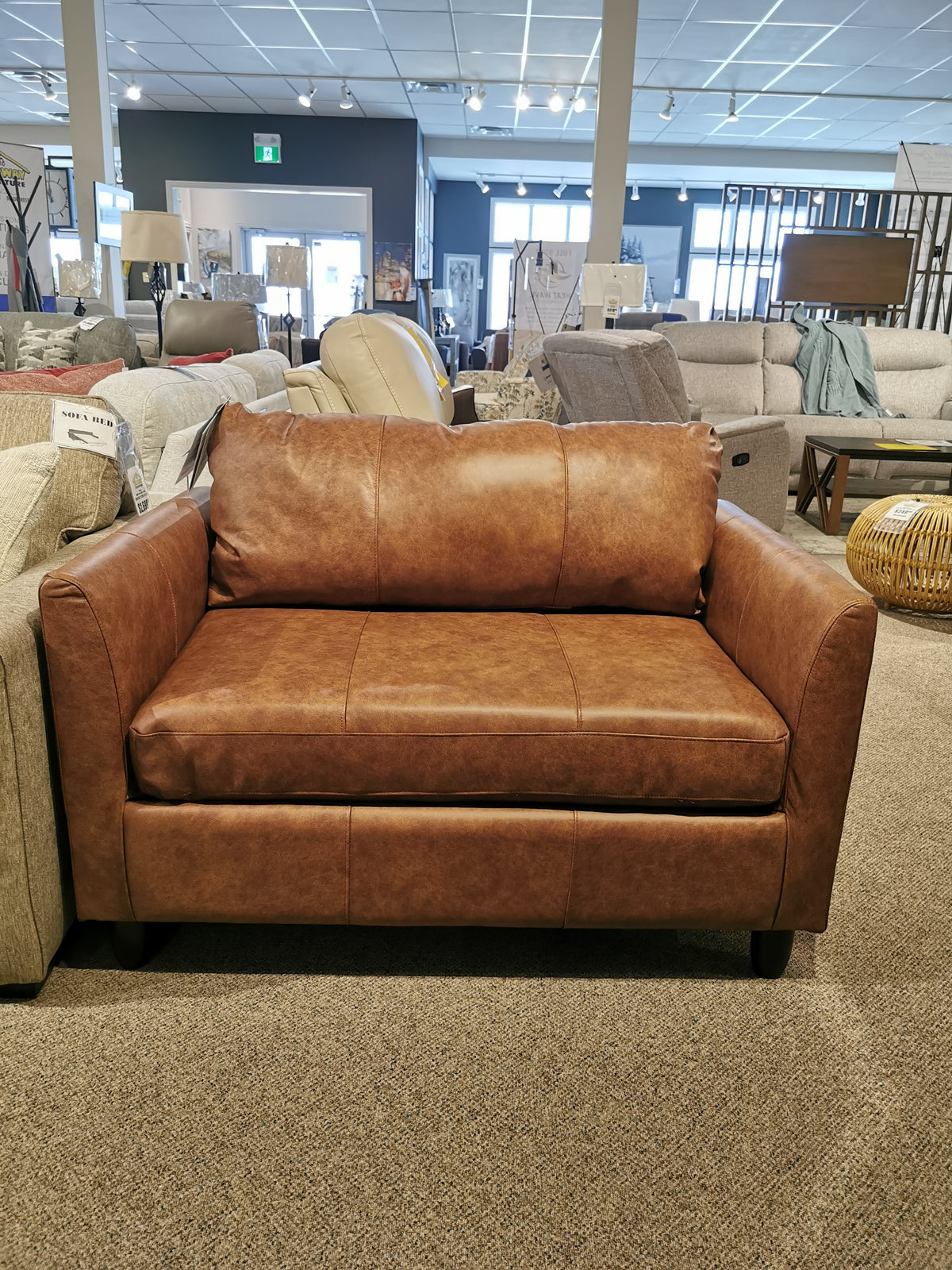 The Bayment Chair Sofa Bed by Best, a brown leather loveseat with sleeper options, is on display in the showroom.