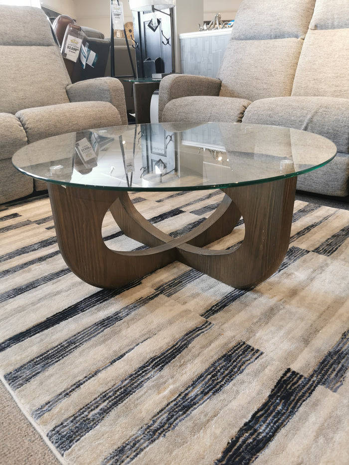 T5951 Lainey Cocktail Table by Magnussen, with a glass top and curved wooden base, on a striped rug between two beige armchairs.