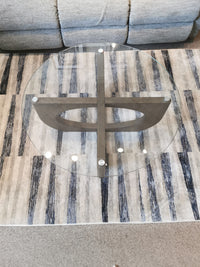 The Magnussen T5951 Lainey table features a round glass top and a cross-shaped pine base on a patterned rug.