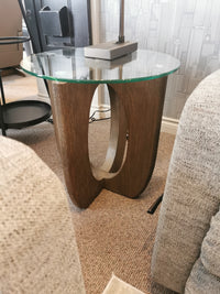 The T5951 Lainey End Table by Magnussen is a stylish glass-top table with a Pine Solids base, perfect between beige sofas.