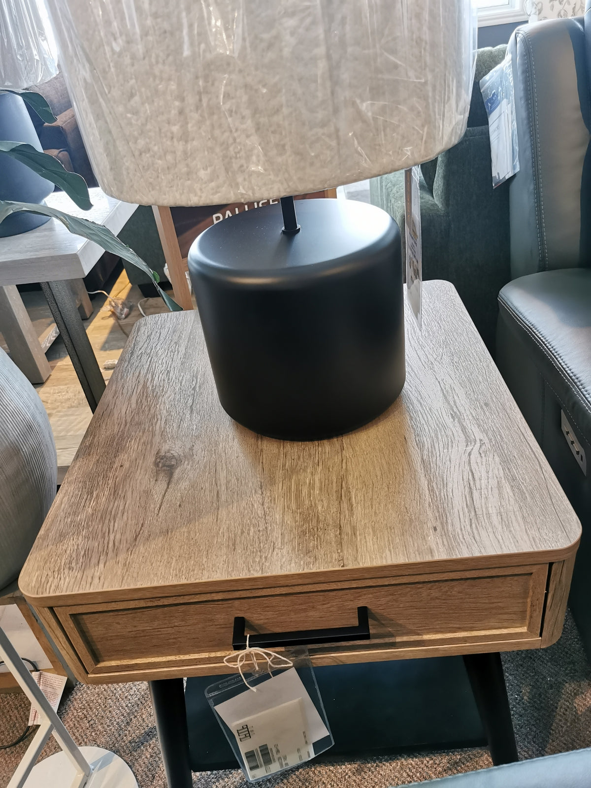 The T5864 Fletcher End Table by Magnussen in Driftwood Finish with a black lamp enhances contemporary charm in the store.