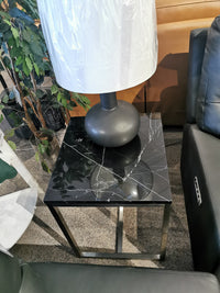 The Magnussen T5760 Kira End Table, a natural black marble top with brushed nickel frame, sits between leather sofas with a ceramic lamp.