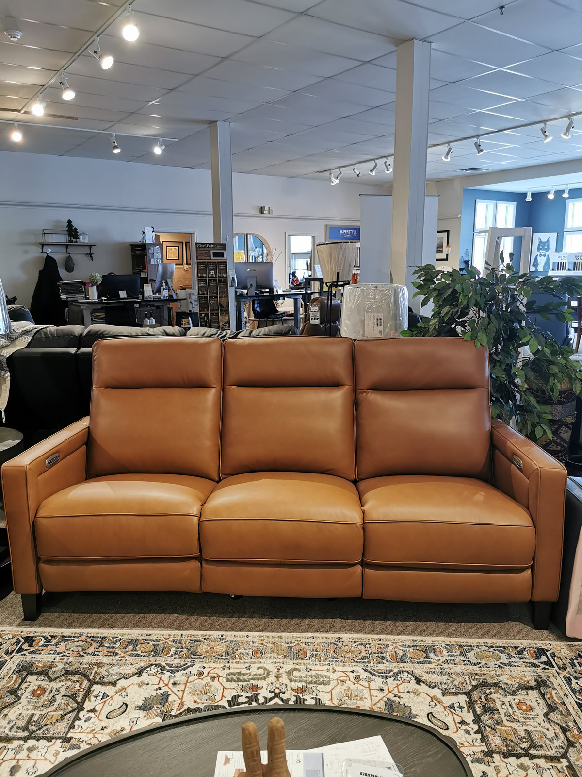 Aurora Furniture's Timberland Power Sofa in brown leather sits on a patterned rug among stylish showroom pieces.