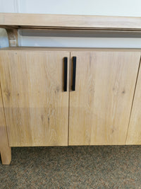 The Somerset 64" Sideboard by Magnussen in butternut with graphite hardware stands on a carpeted floor and white wall.