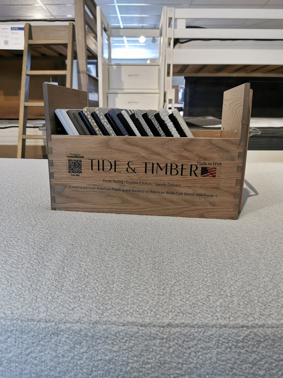 A wooden box labeled "Tide & Timber" displays solid oak samples on a mattress in a showroom with Vaughan Bassett daybeds.