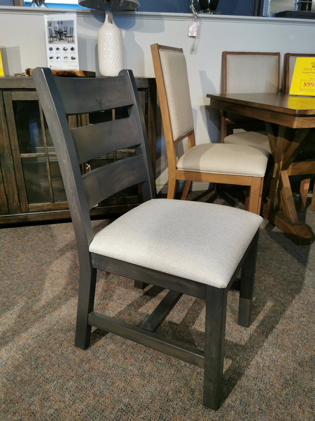 Wooden chair with gray cushion next to a Handstone Parker 5pc Dining Package in a showroom.