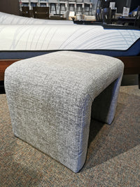 The Sophia Petite Bench by Jofran, in gray luxe chenille fabric, is on a carpeted floor by a bed with a white mattress.