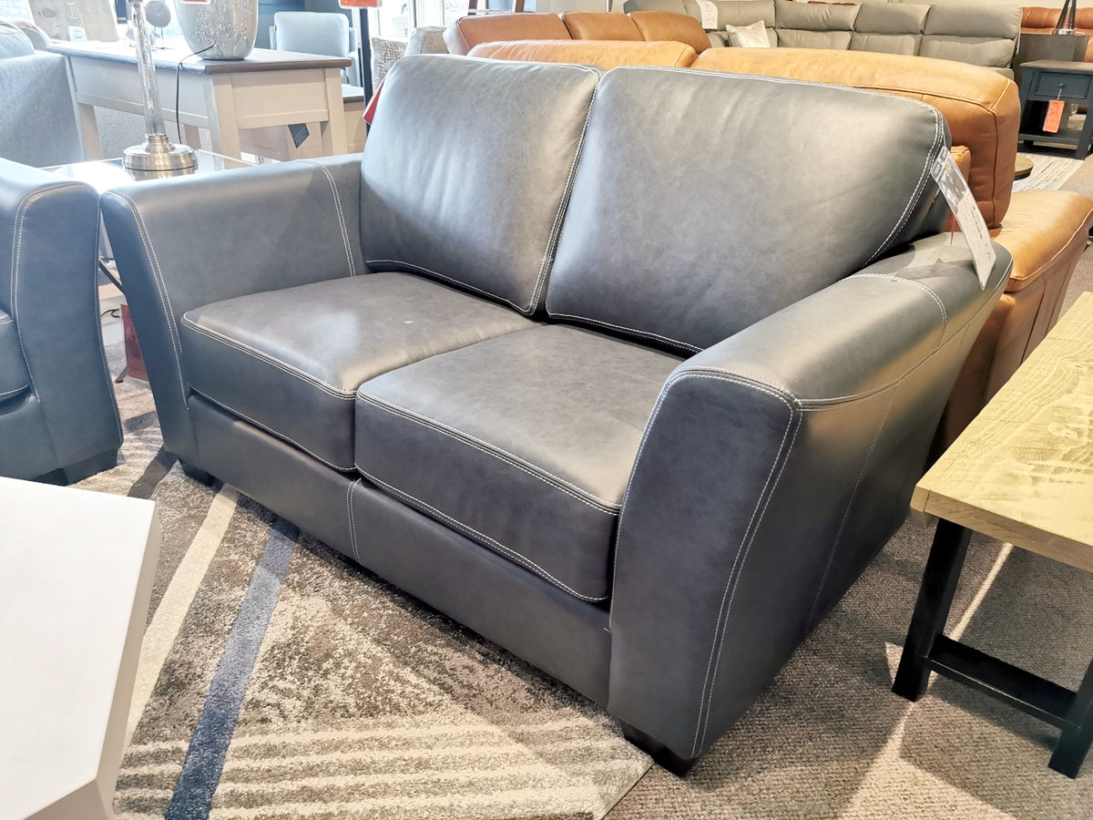 The Bayview Leather Loveseat from Leather Craft, a timeless dark gray leather loveseat featuring wide armrests, is displayed in a furniture store showroom.