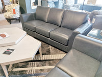 Modern contemporary living room featuring a Bayview Leather Sofa by Leather Craft in gray leather, complemented by a sleek coffee table and patterned rug, with additional upholstered furniture surrounding the space.